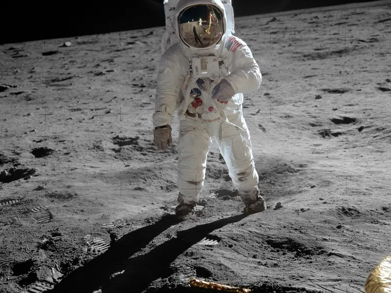 An astronaut in a white spacesuit standing on grey ground, the gold visor reflecting the photographer and the lander