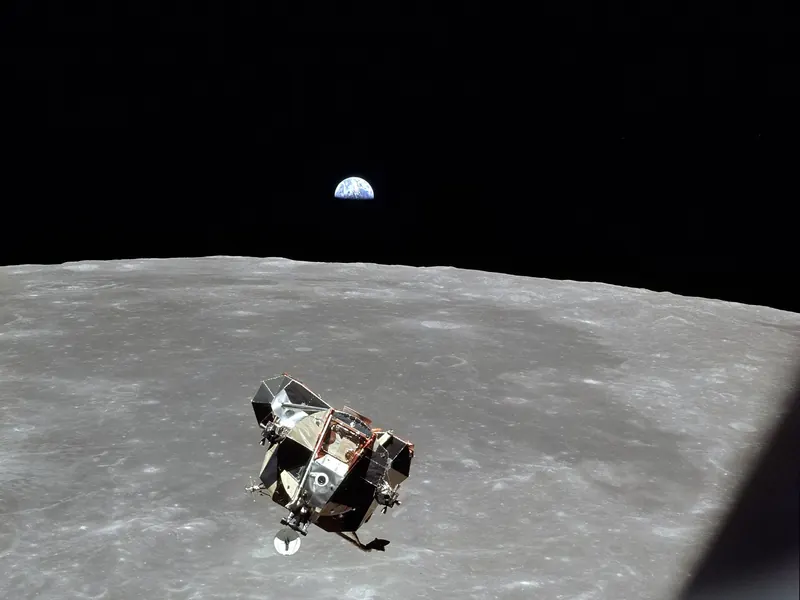 A spidery lander with gold foil flying above the grey Moon, with a small Earth on the horizon
