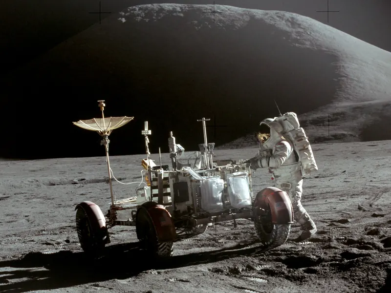 An astronaut beside a four-wheeled lunar rover with a dish antenna, with a mountain behind