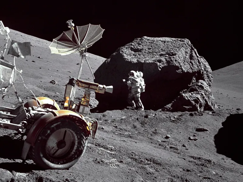 An astronaut standing next to a huge split boulder, with the lunar rover in front